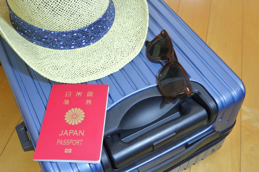 A red Japan passport resting on a blue suitcase with a straw hat and sunglasses nearby on a wooden floor.