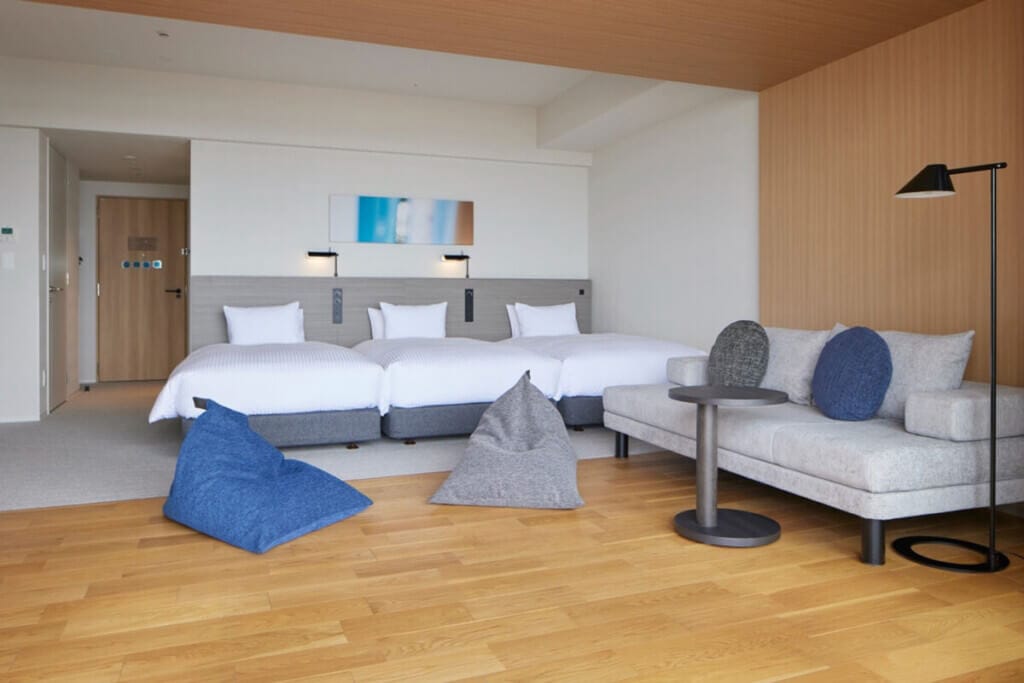 Hotel room with three single beds, white bedding, and a gray headboard, plus a seating area with sofa and cushions