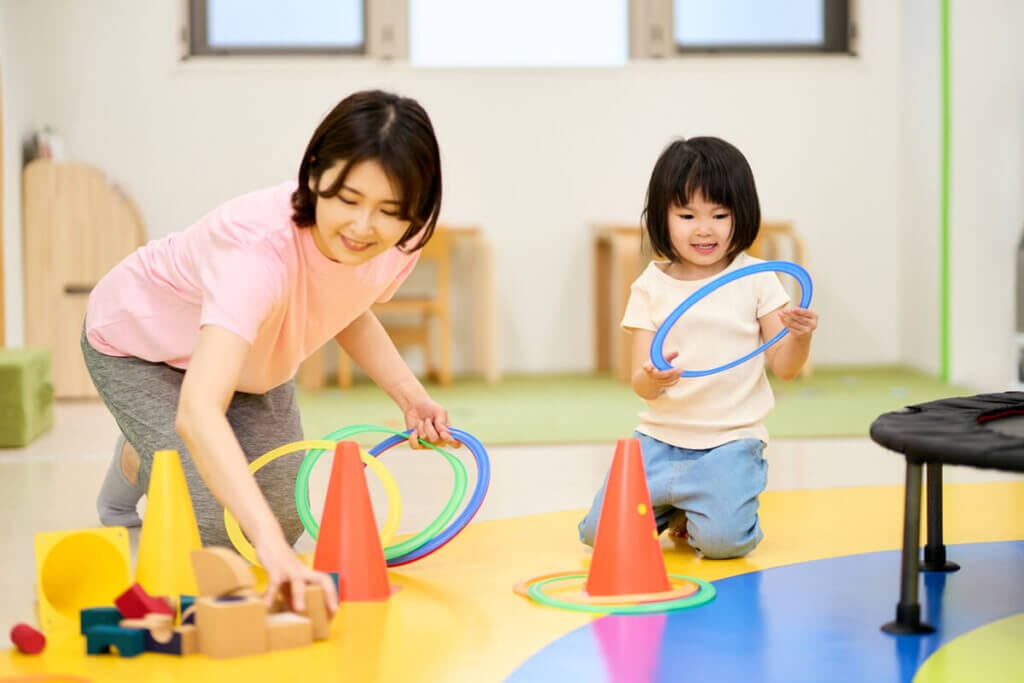 Woman and young girl playing with colorful hoops and cones on a bright yellow play mat in a classroom-like room.
