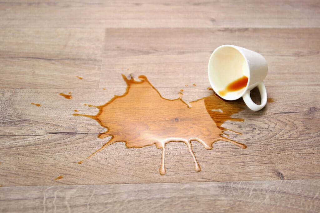 White ceramic mug tipping over with coffee spilled across a wooden floor, forming irregular brown stains