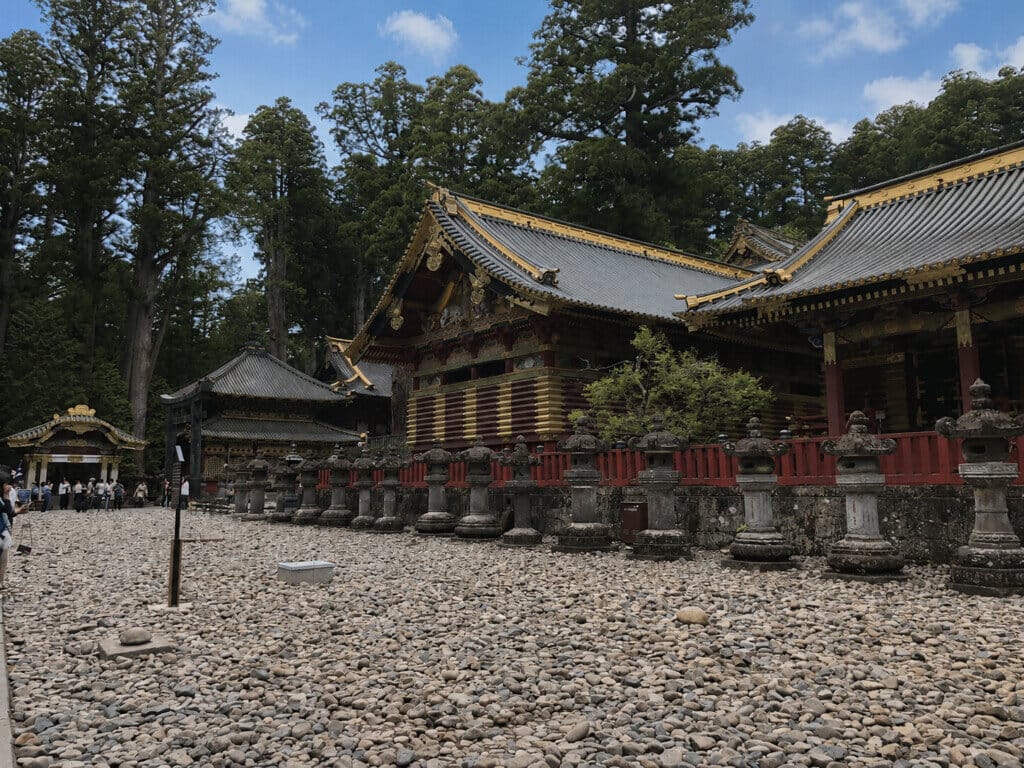 Japanese temple with gilded roof trim and ornate wooden structure, lined by stone lanterns on a pebble courtyard
